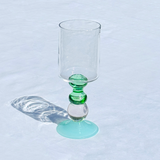 A clear glass goblet with a green stem and a light aqua-colored circular base, photographed on a white textured fabric surface with a soft shadow cast to the left.
