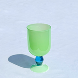 A translucent light green goblet glass with a blue faceted stem, standing on a white textured surface with a soft shadow to the left.