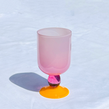 A pink frosted glass goblet with a vibrant orange-yellow circular base, photographed on a white textured fabric surface.
