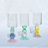 Three clear glass candle holders with colorful, bulbous bases in pink, blue, green, white, yellow, and purple, arranged on a white fabric background.