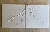 Two square ceramic tiles with embossed leafy branch patterns, displayed side-by-side on a rustic wooden surface.