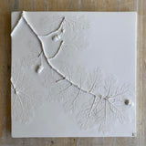 A square white decorative tile featuring embossed oak leaves and branches, displayed flat on a rustic, light brown wooden surface.