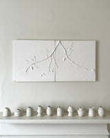 A white two-panel wall art piece with a raised branch and bead motif, displayed above a white mantel with a row of small speckled ceramic vases against a light beige wall.