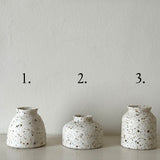 Three small speckled ceramic vases in different heights are lined up on a white shelf against a textured off-white wall, with black numbers 1, 2, and 3 above each vase.