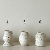 Three small speckled ceramic vases in off-white with brown and tan flecks, displayed on a white shelf against a light wall, labeled 4, 5, and 6 above each vase.
