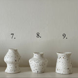 Three small speckled ceramic vases in different bulbous shapes are lined up on a white shelf against a textured beige wall, with black numbers 7, 8, and 9 printed above each vase.