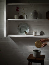 A rustic interior scene with white shelves holding ceramic vases, a plate, and small vessels against a white brick wall. A person's hands are visible at the lower right holding a round wooden object above a gray ceramic pot on a small wooden stool.