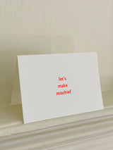 A white A5 size greeting card with the phrase 'let's make mischief' printed in red lowercase letters, displayed standing on a white mantelpiece against a beige wall.