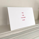 A white folding letterpress greeting card with red text that reads 'my cup runneth over' displayed on a table.